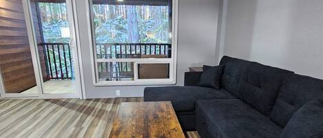Un salon moderne avec un canapé d’angle gris foncé, une table basse en bois et une grande porte-fenêtre coulissante donnant sur un balcon avec vue sur les arbres.