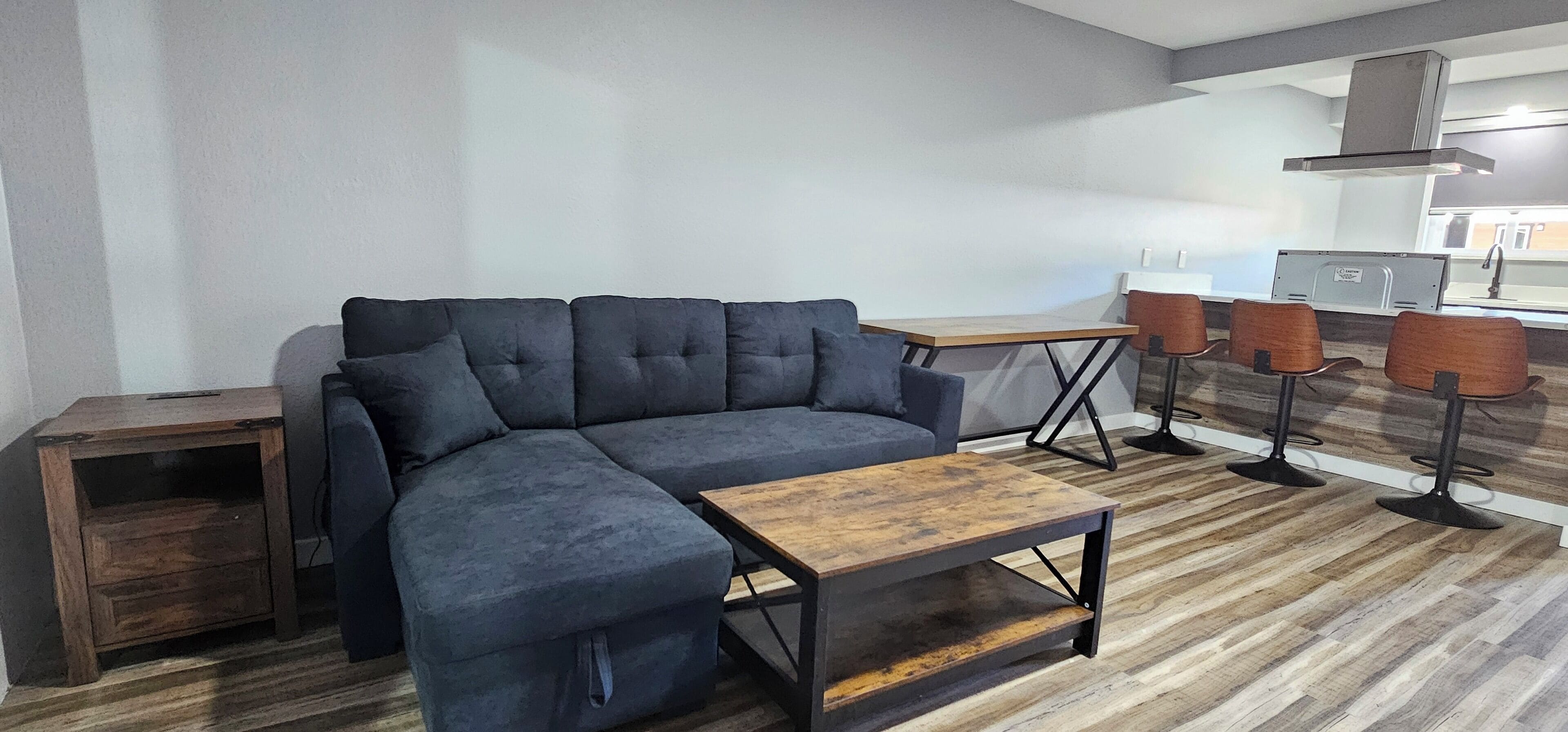 Een moderne woonkamer met een hoekbank, een houten salontafel en een keukenhoek met barkrukken.