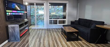 Una sala de estar moderna con un televisor de pantalla plana, un sofá oscuro, una mesa de centro de madera y un ventanal corredizo que da a un balcón.