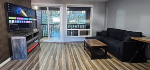 A modern living room with a flat-screen TV, a dark sofa, a wooden coffee table, and a sliding glass door leading to a balcony.
