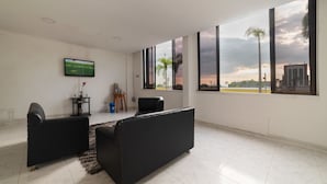 Una sala moderna con televisor de pantalla plana, dos sofás negros, una mesita auxiliar y grandes ventanas con vista a palmeras y un atardecer.