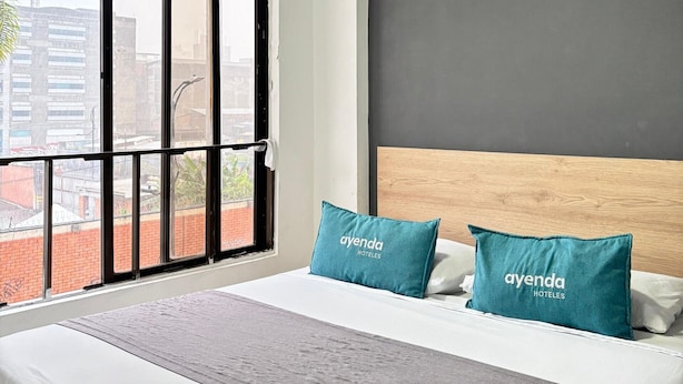 Habitación de hotel con una cama grande, dos almohadas con la marca 'ayenda hotels', una colcha gris y vistas de edificios a través de un ventanal.