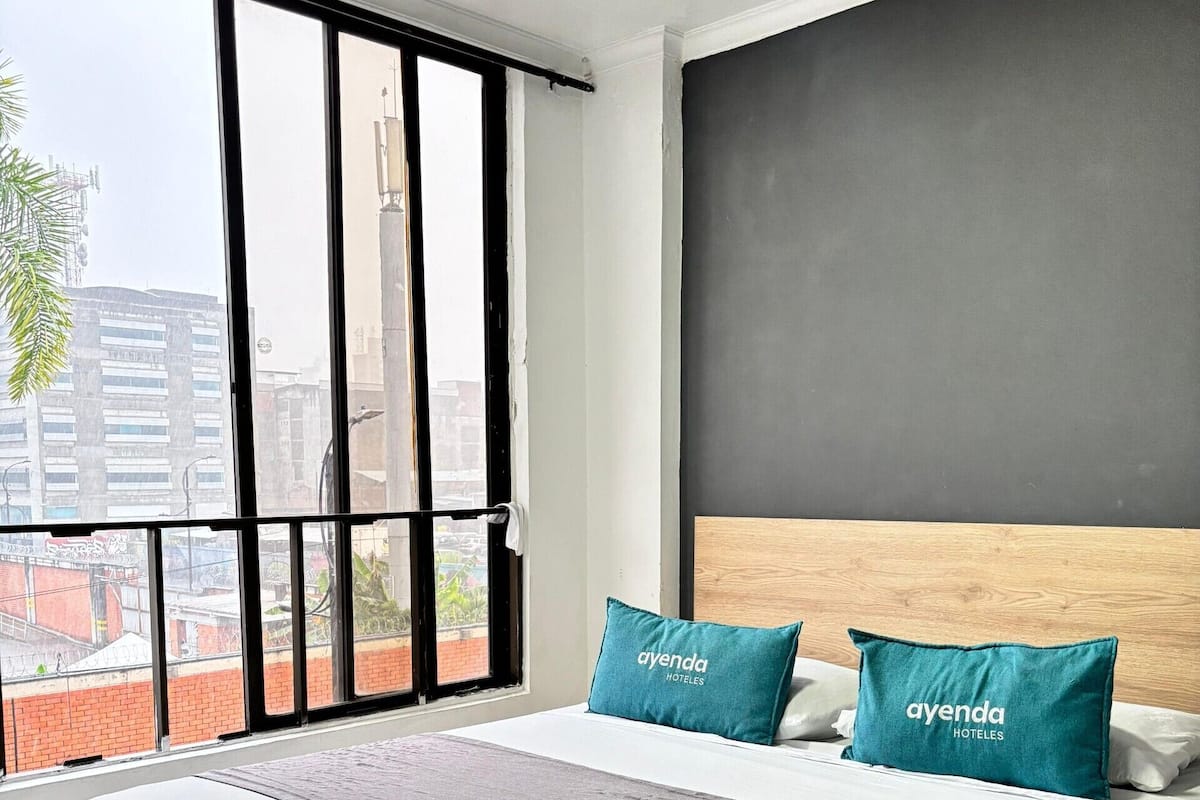 A hotel room with a large bed, two pillows with the hotel name, a grey throw, and a view of buildings through a window.
