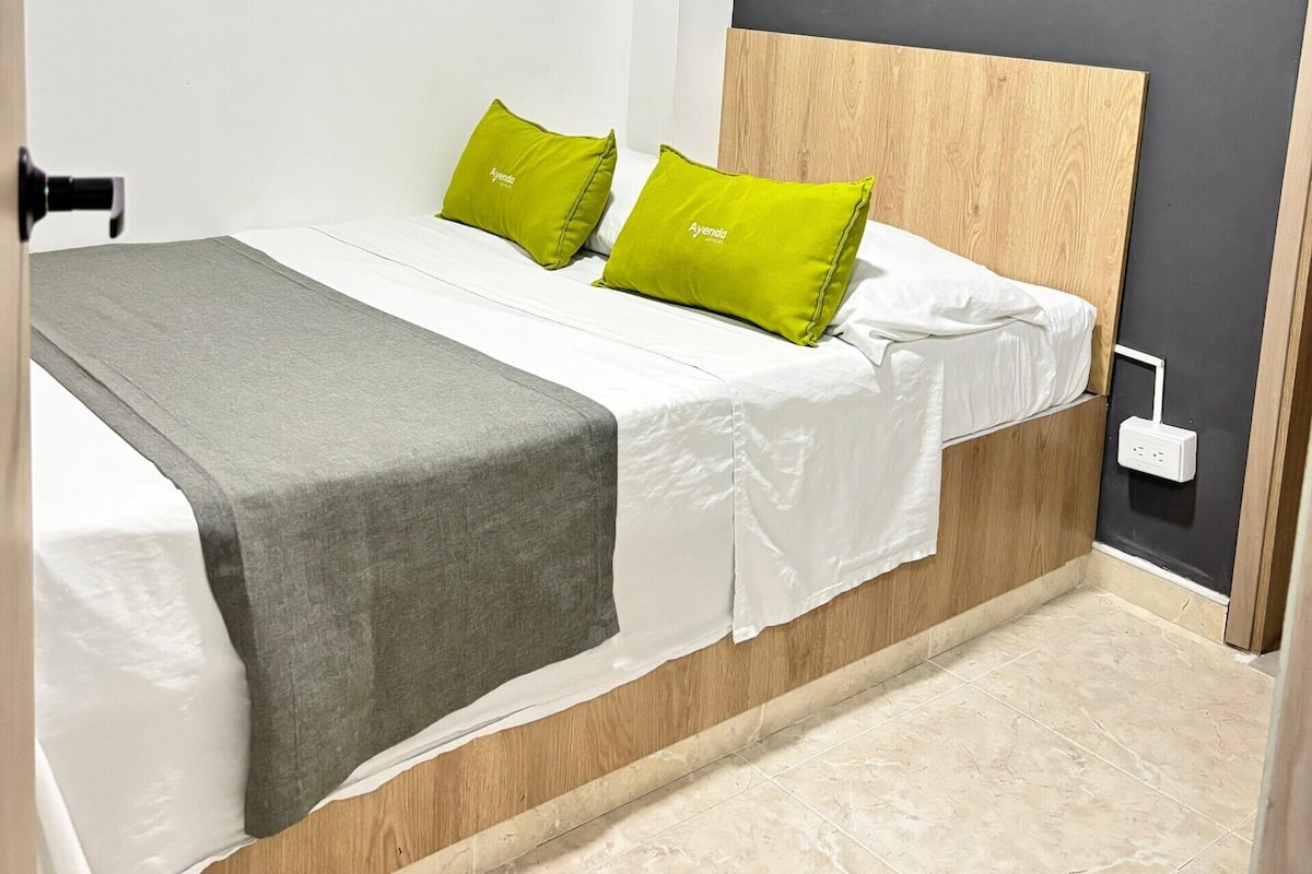 A modern bedroom with a bed, two green pillows, a grey blanket, and a wooden headboard.