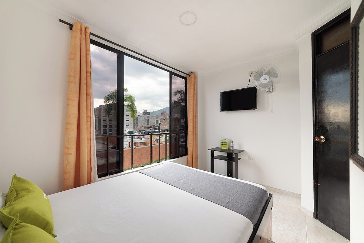A hotel room with a bed, a small table with a bottle and a box, a TV on the wall, and a door.