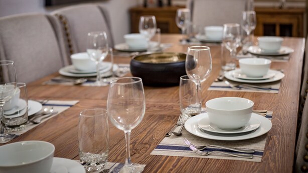 Mesa de jantar posta com pratos, tigelas e copos de vinho brancos, sobre mesa de madeira e jogos americanos.