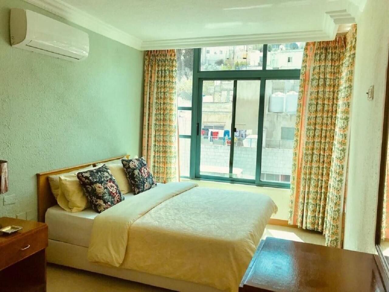 Ein Schlafzimmer mit einem Bett, einem Nachttisch, einem Fenster mit Vorhängen und einer Klimaanlage.