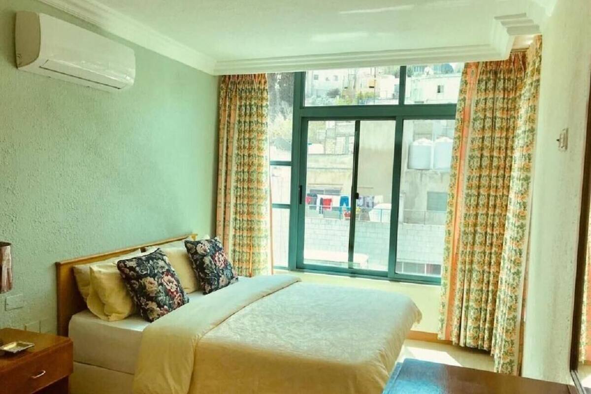 A bedroom with a bed, a nightstand, a window with curtains, and an air conditioning unit.