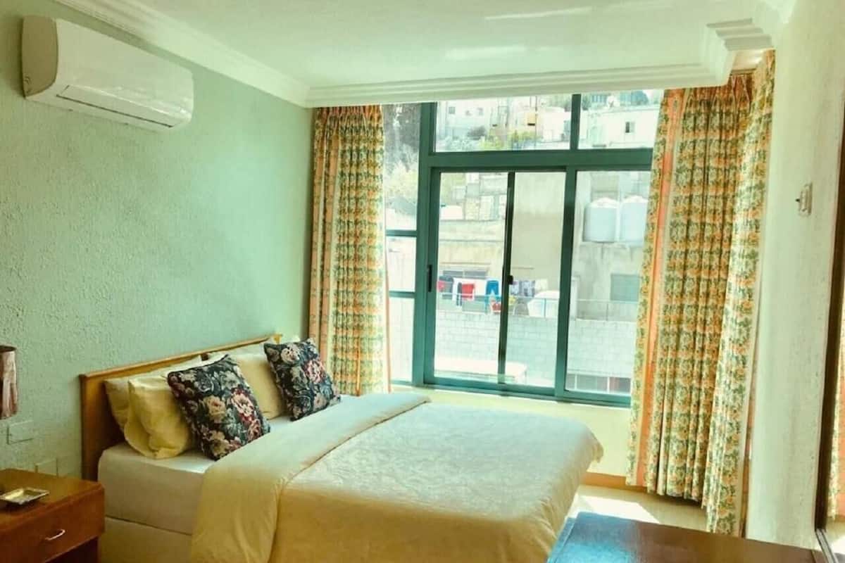 Ein Schlafzimmer mit einem Bett, einem Nachttisch, einem Fenster mit Vorhängen und einer Klimaanlage.