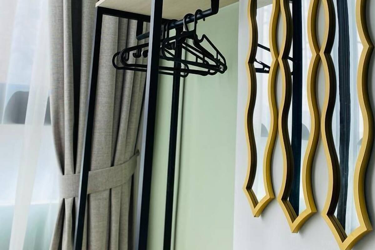 A room with a window, a metal drying rack, and a wall-mounted mirror with a wavy frame.