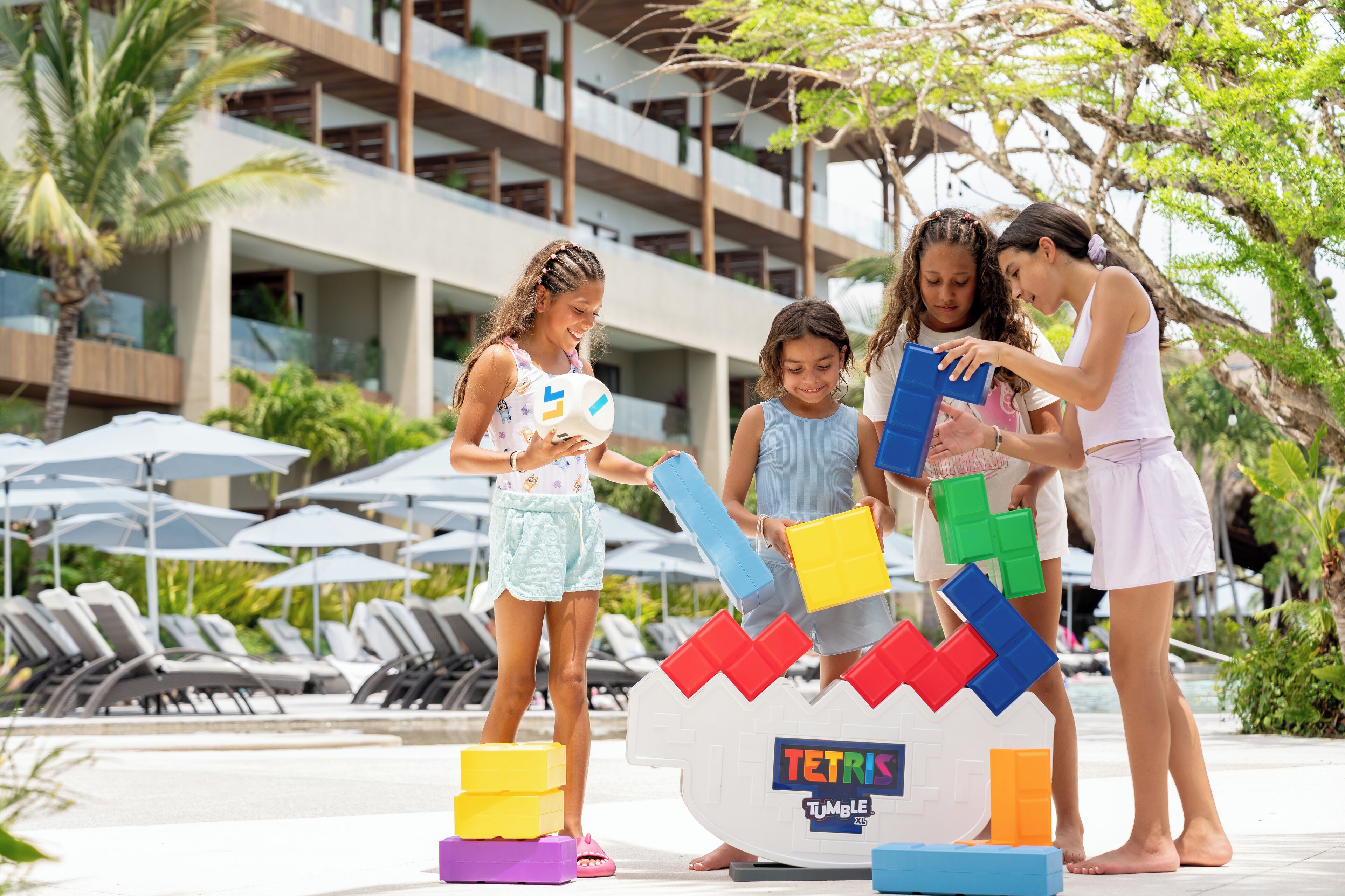 Eine Gruppe Kinder spielt mit bunten Bauklötzen in der Nähe eines Poolbereichs mit Liegestühlen und Sonnenschirmen.