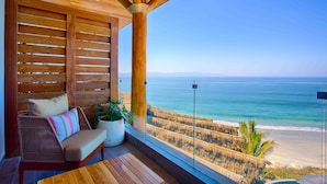 Une terrasse en bois avec un canapé en osier et un pot de fleurs, donnant sur une plage et offrant une vue dégagée sur l’océan.