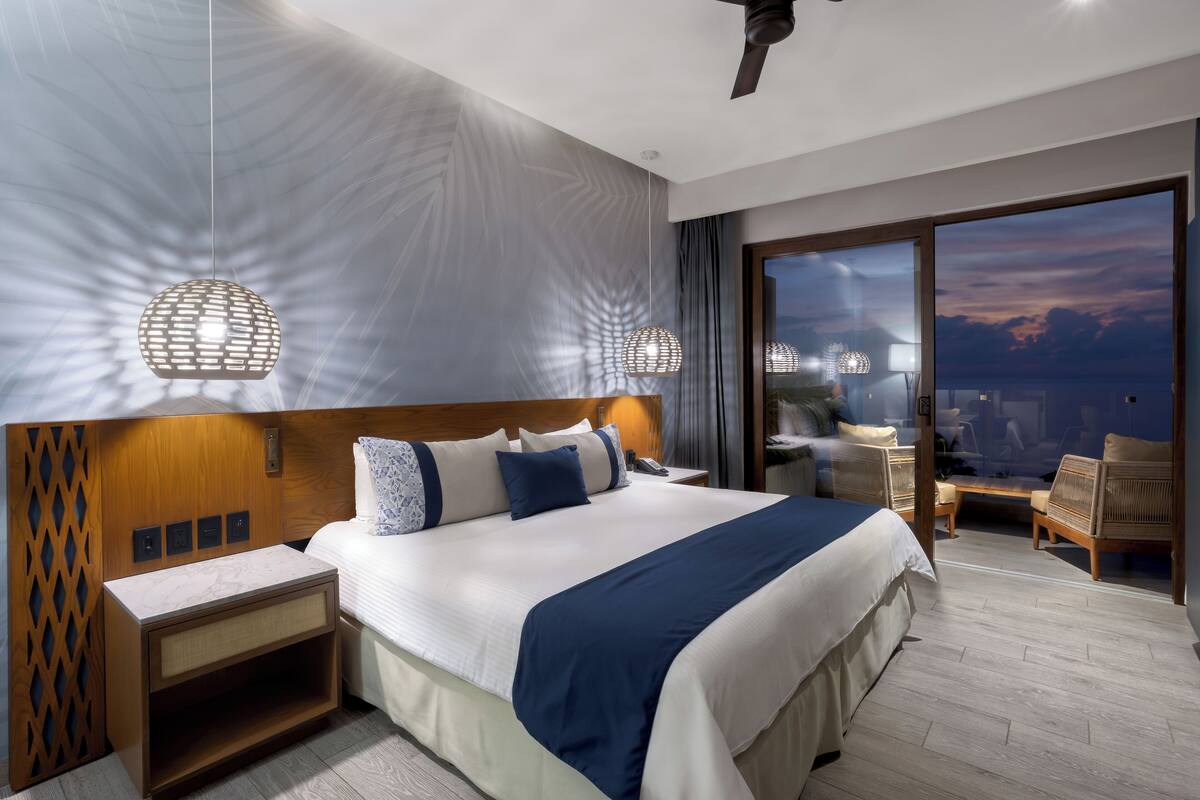 Een moderne hotelkamer met een groot bed, een houten hoofdbord, een nachtkastje en uitzicht op de stad door een groot raam.