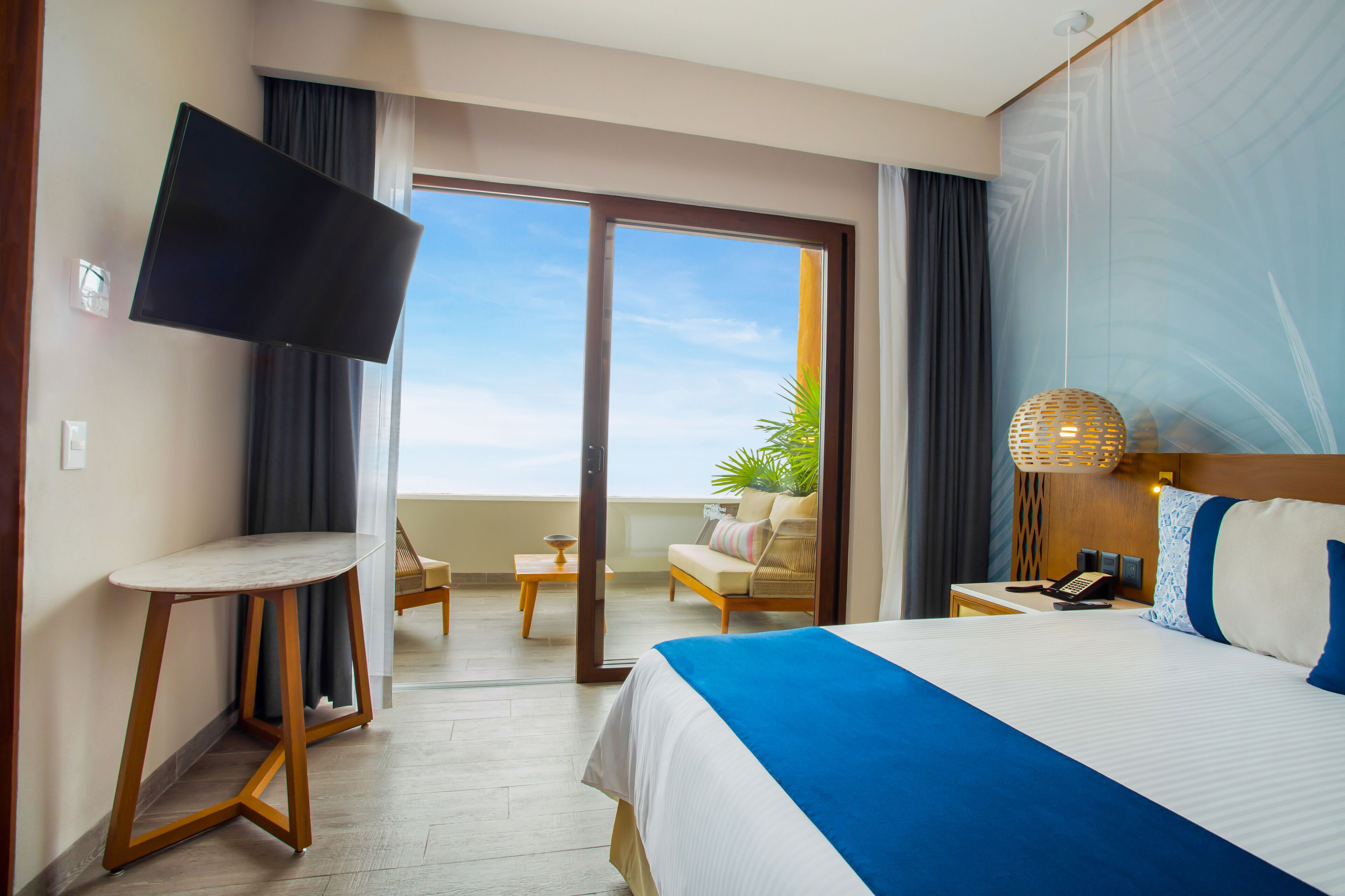 A hotel room with a bed, a TV, a small table, a chair, a lamp, and a view of the outdoors.