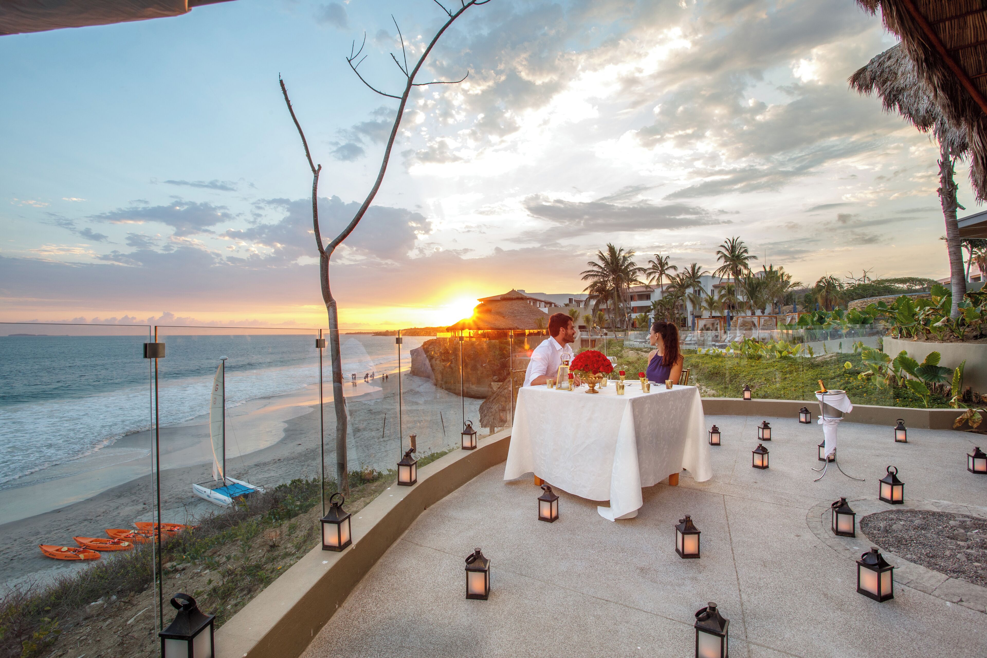 A sunset view from a terrace with a dining table set for two, overlooking the beach and ocean.