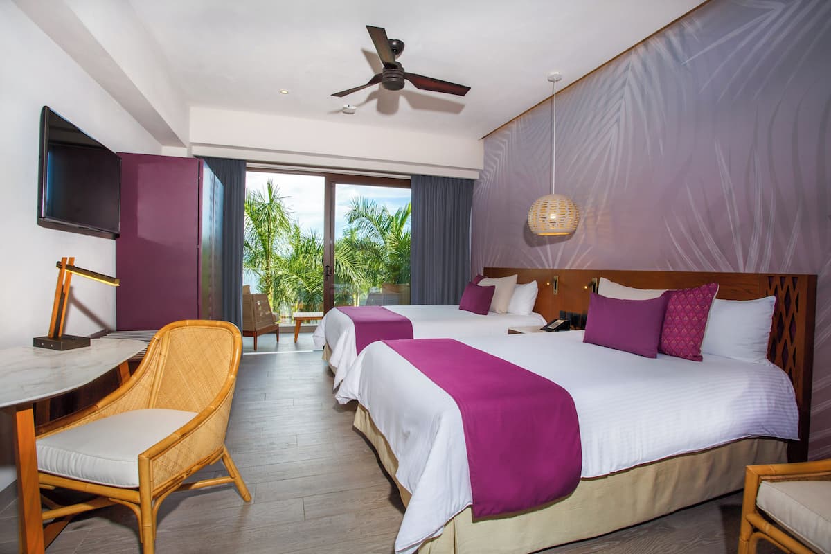 Hotelkamer met een groot bed, twee nachtkastjes, een bureau, een stoel, een plafondventilator en een groot raam met uitzicht op palmbomen.