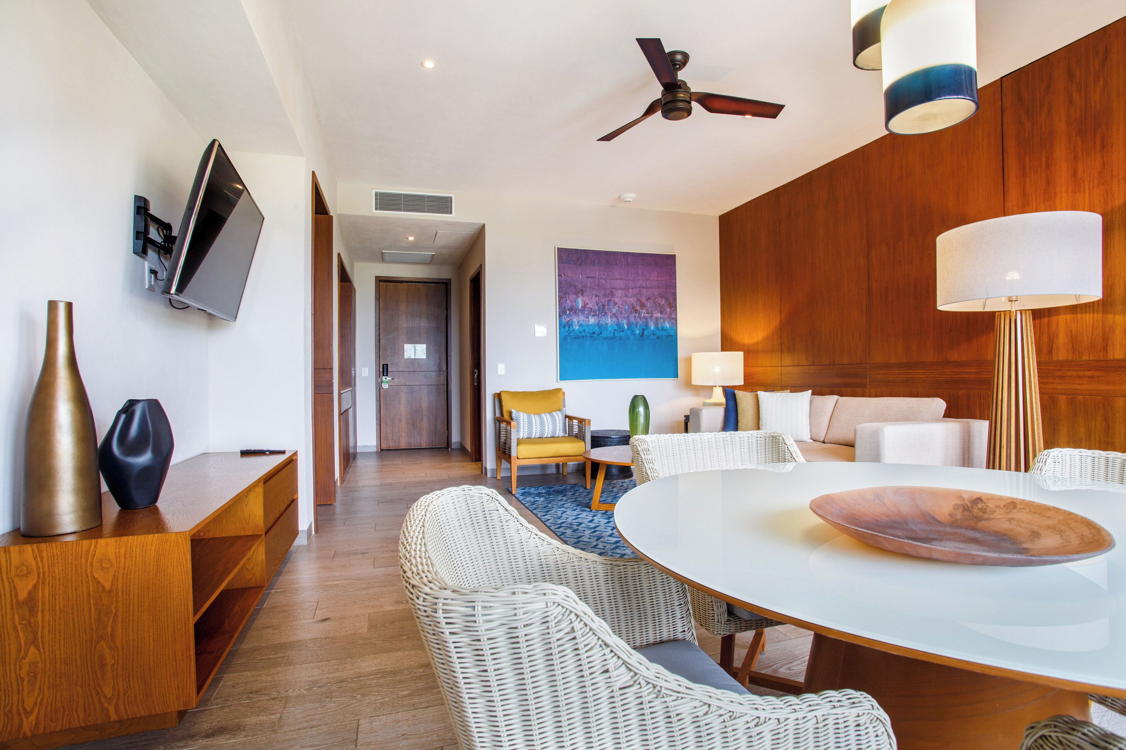A modern hotel room with a dining area, a flat-screen TV mounted on the wall, a ceiling fan, and a large painting on the wall.