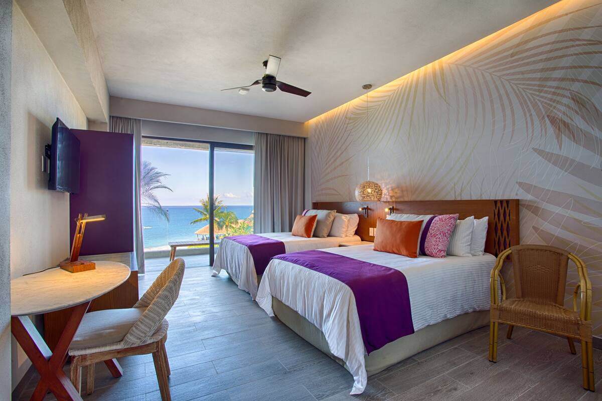 Een hotelkamer met twee bedden, een bureau, een stoel, een plafondventilator en uitzicht op de oceaan.