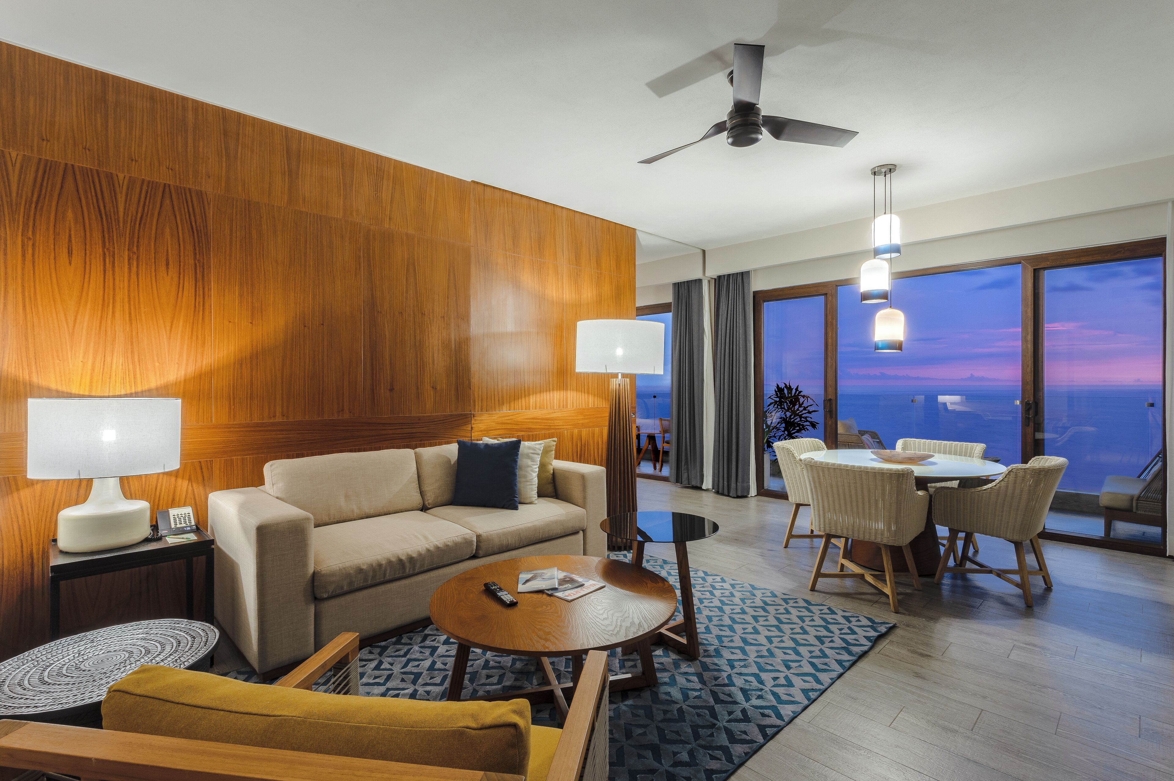 Ein modernes Hotelzimmer mit einer Wand in Holzoptik, einer Couch, einem Couchtisch, einem Essbereich mit Tisch und Stühlen sowie großen Fenstern mit Blick.