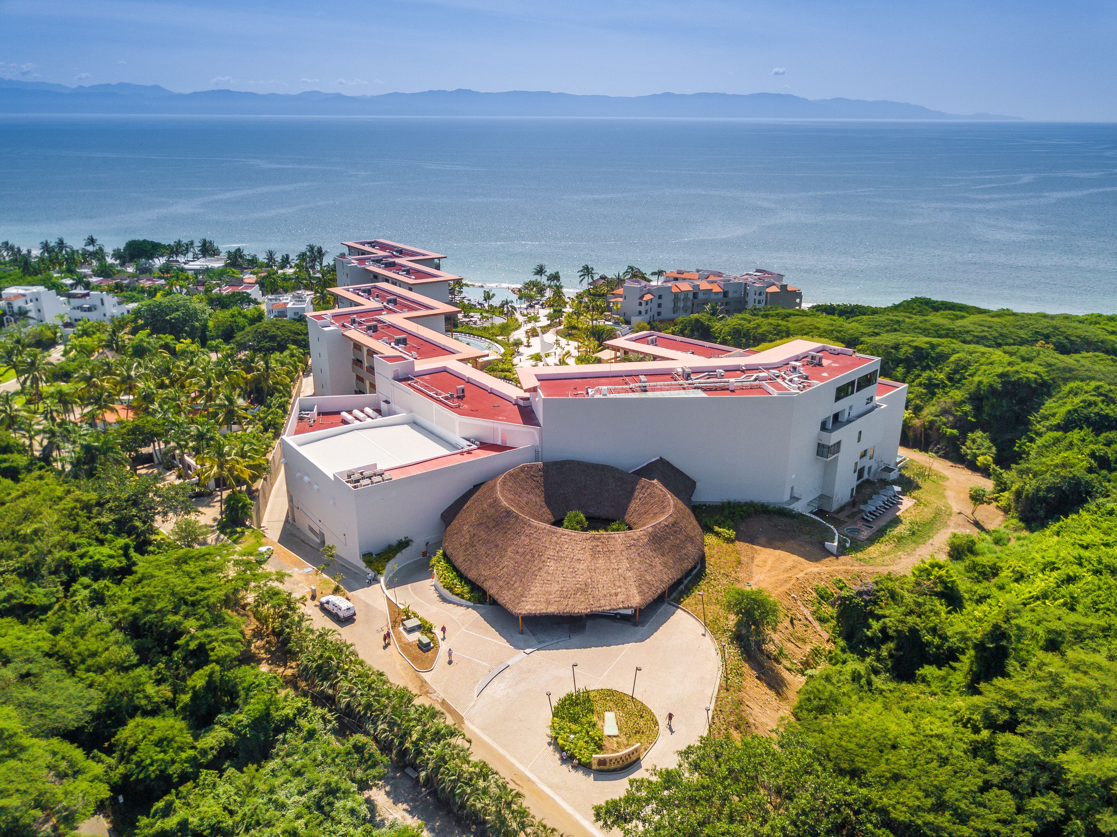 A modern hotel with a unique circular structure, surrounded by lush greenery and a view of the sea.