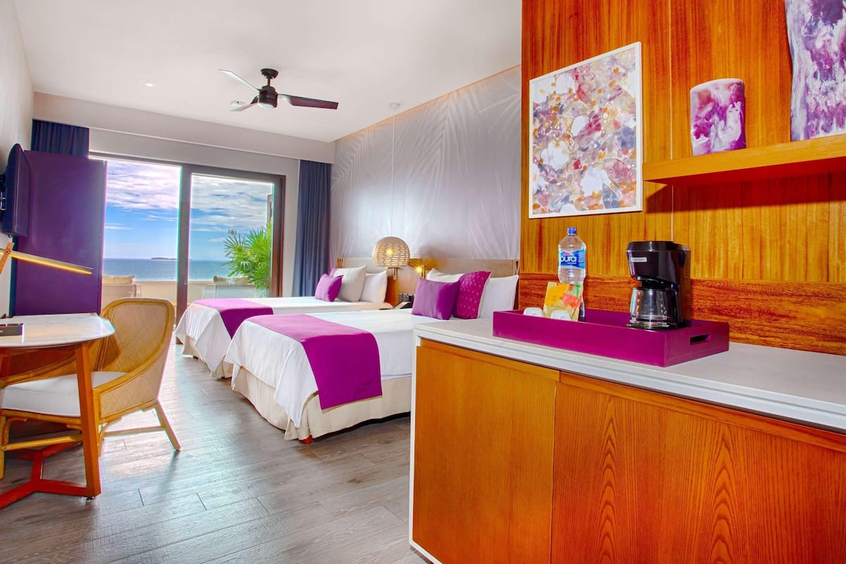 Een hotelkamer met twee bedden, een bureau, een stoel, een koffiezetapparaat en kunst aan de muur.