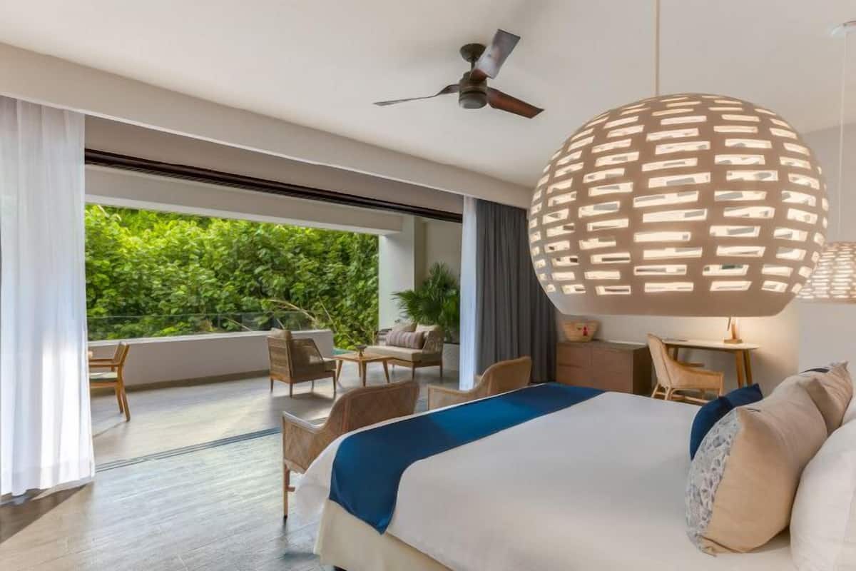 Een moderne hotelkamer met een groot bed, een plafondventilator, een balkon met uitzicht op groen en een opvallende, sferische lamp.