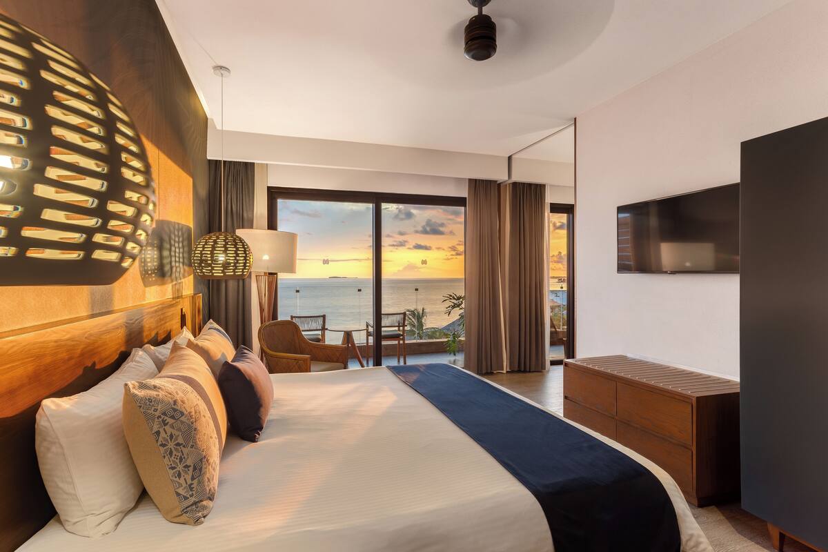 Een hotelkamer met een groot bed, een televisie, een balkon met uitzicht op zee en een plafondventilator.