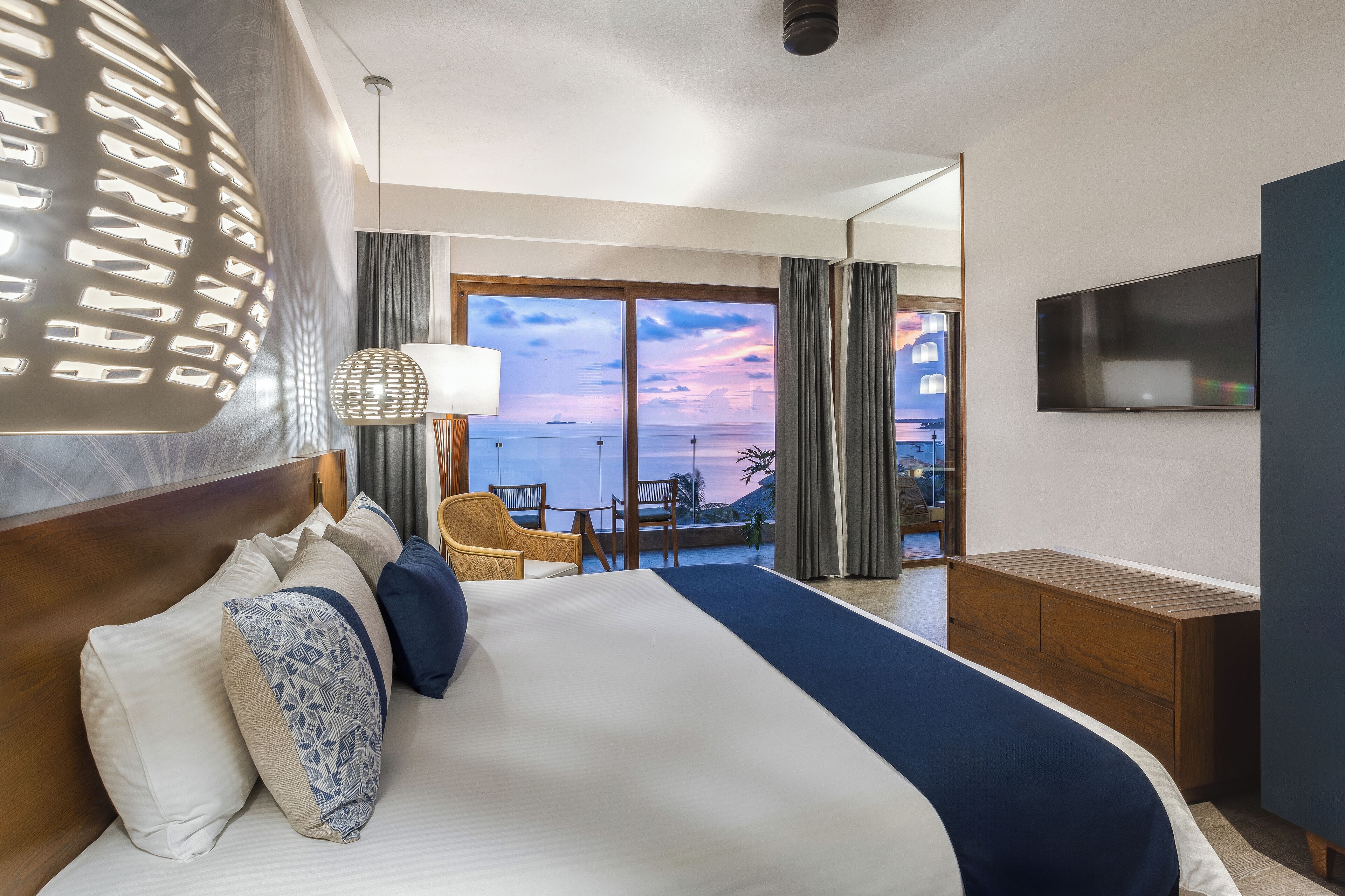 Ein Hotelzimmer mit einem großen Bett, einem Fernseher, einem Schreibtisch und einem Balkon mit Meerblick.