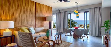 A modern hotel room with a wooden accent wall, a sofa, a dining table with chairs, a ceiling fan, and a view of the ocean.