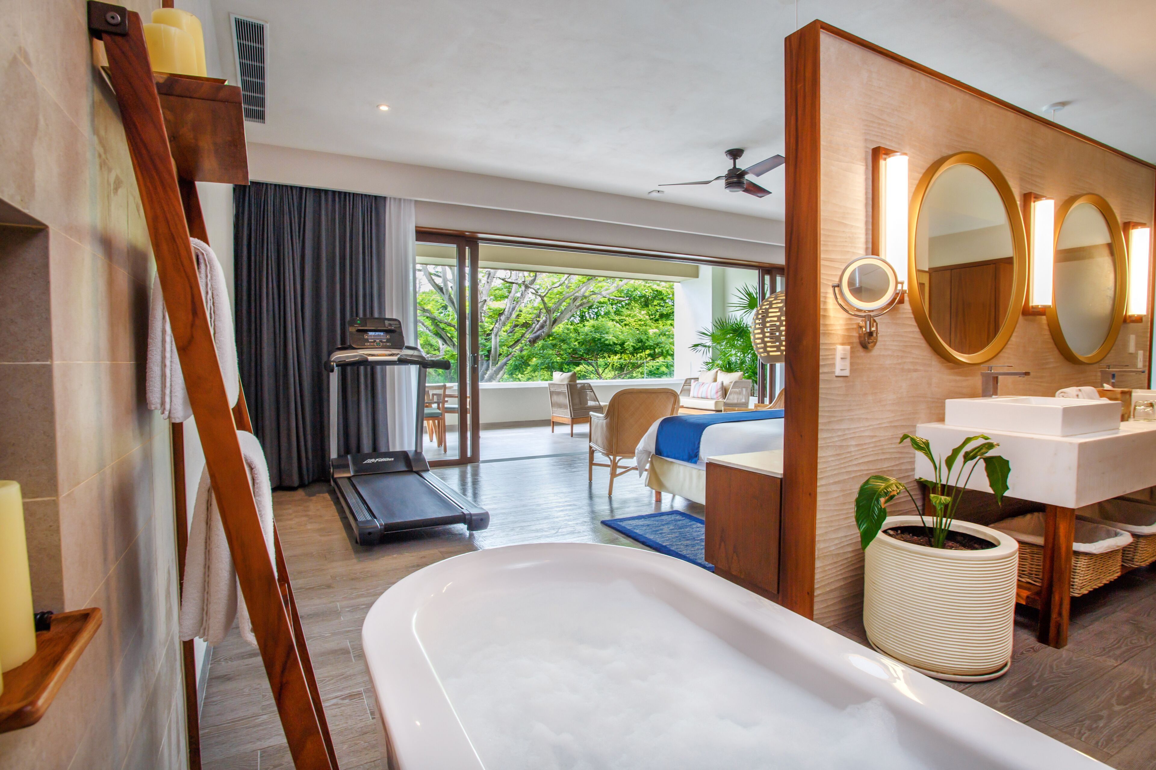 Une salle de bain moderne équipée d’une baignoire, d’un tapis de course et d’une grande fenêtre donnant sur un paysage verdoyant.