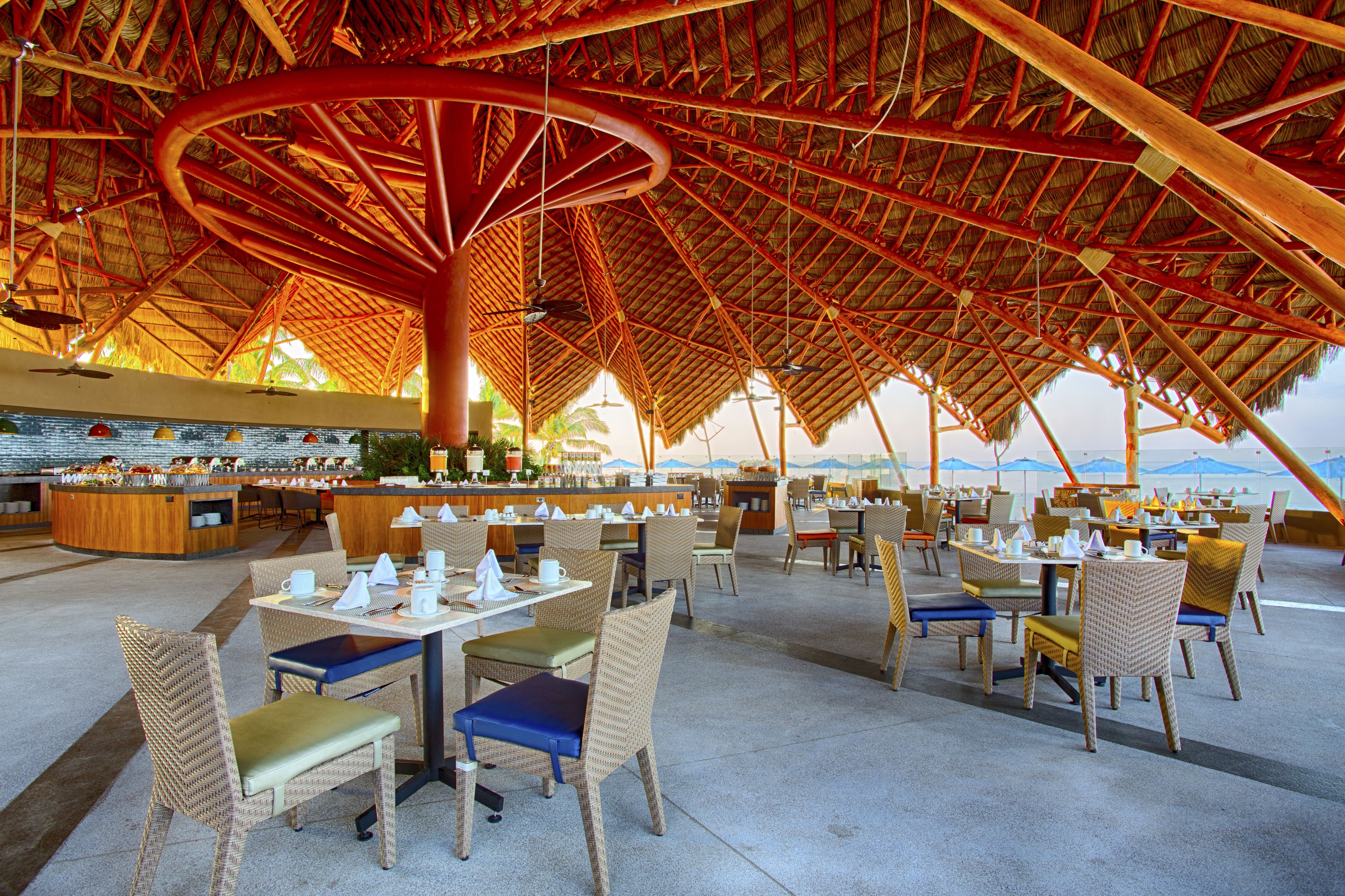 Un área de comedor al aire libre con sillas y mesas de mimbre, una barra central y una estructura de techo de paja.