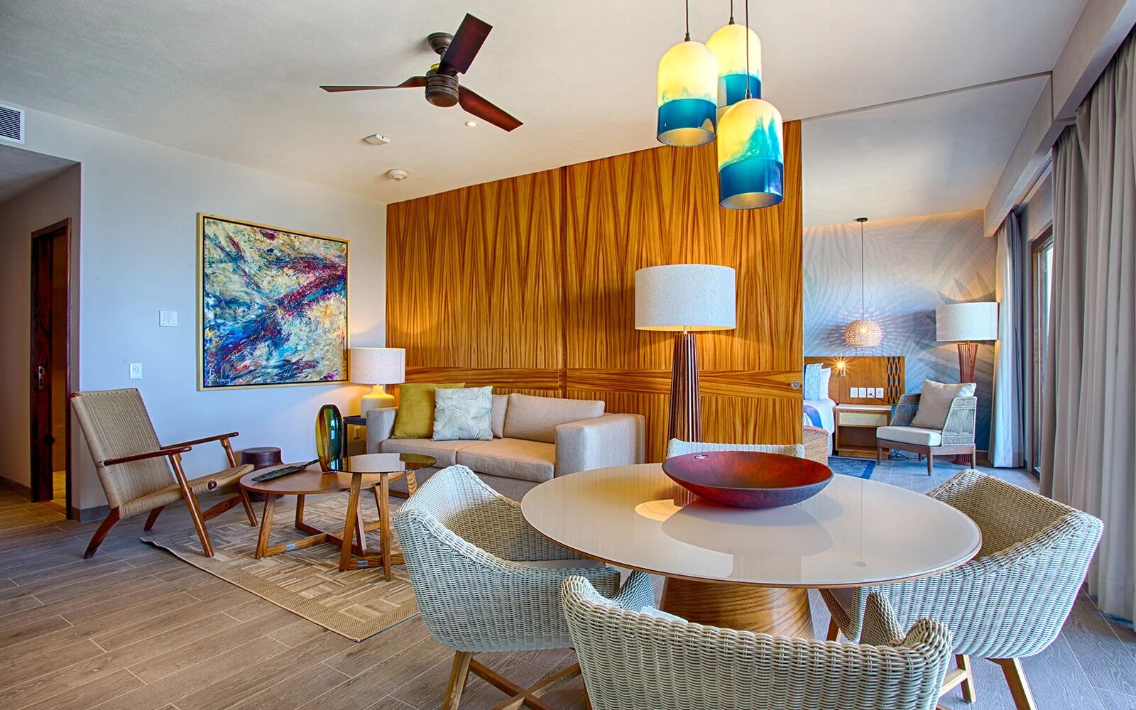 A modern hotel room with a round dining table, wicker chairs, a sofa, and a ceiling fan.