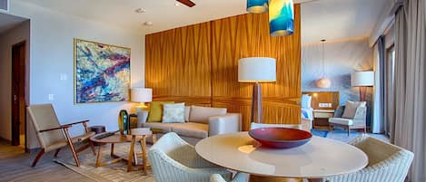 A modern hotel room with a round dining table, wicker chairs, a sofa, and a ceiling fan.