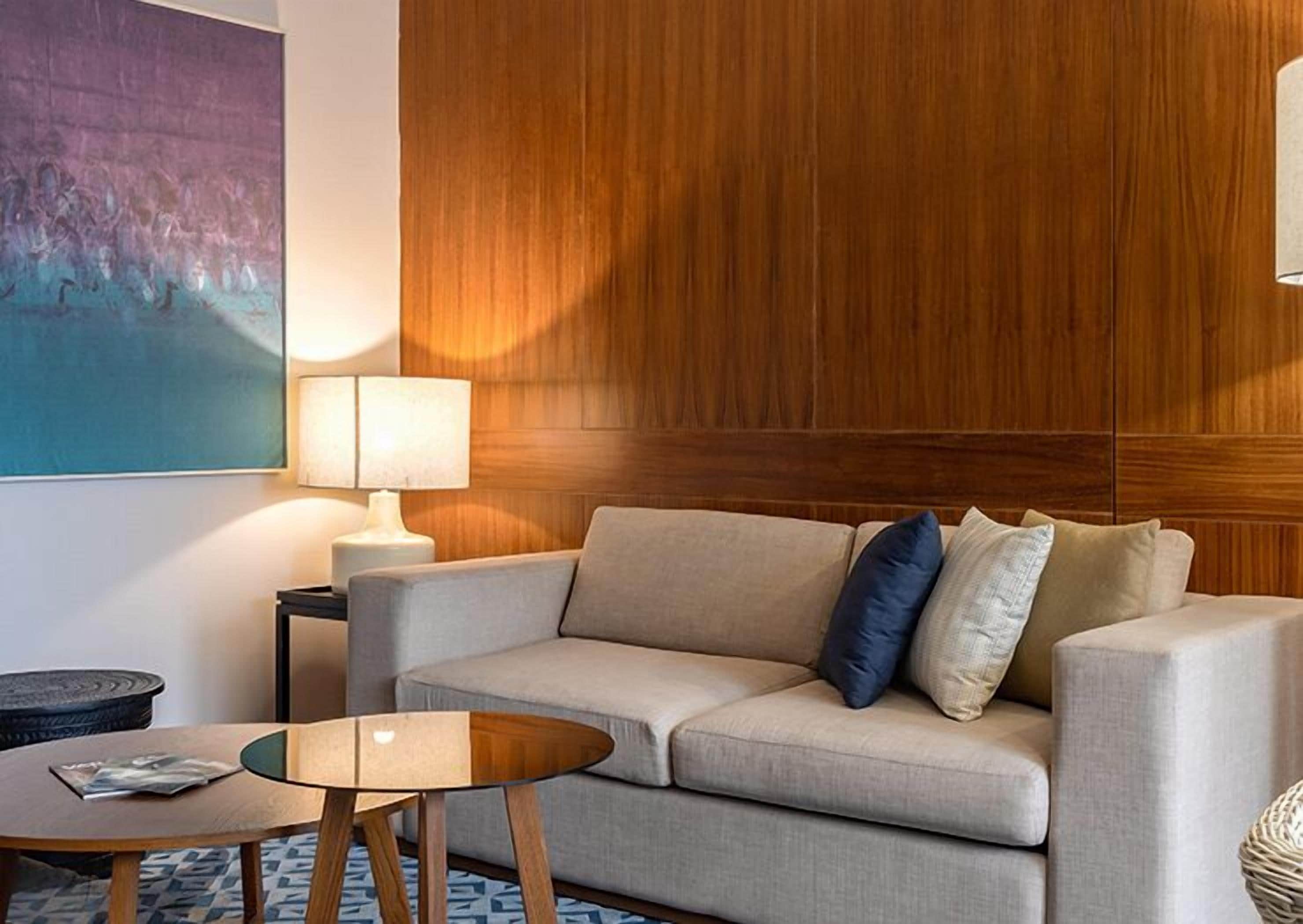 A modern living room with a beige sofa, a glass coffee table, a lamp, and a painting on the wall.