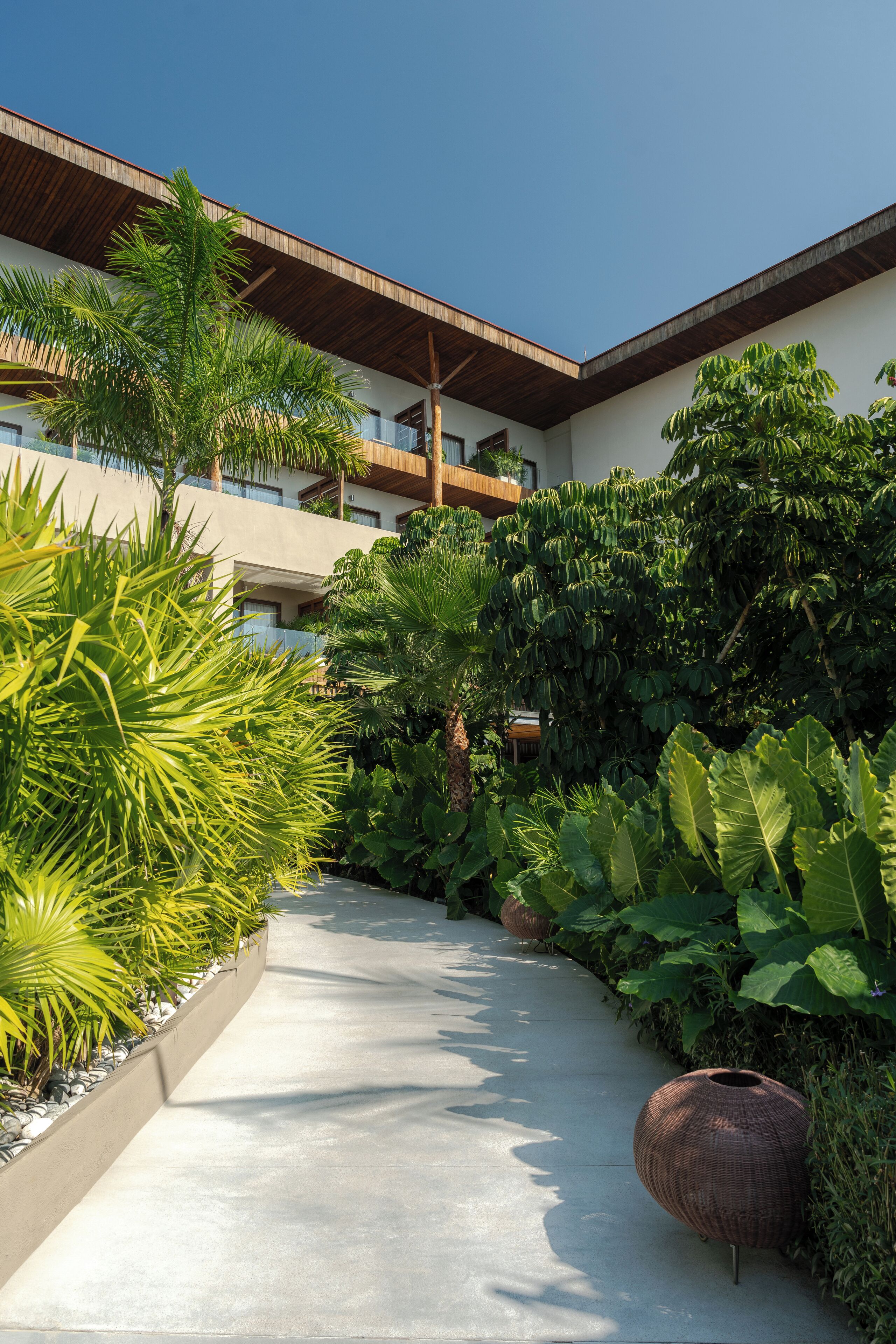 A pathway flanked by lush greenery and a large decorative vase, leading to a building with a wooden roof and multiple balconies.