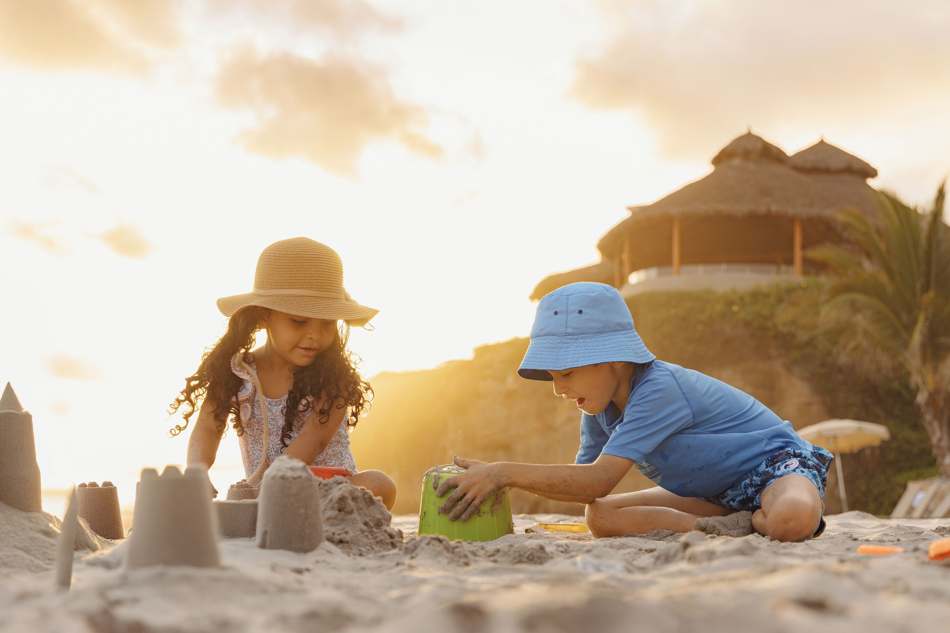 Dos niños construyen castillos de arena en una playa, con una estructura de techo de paja al fondo.