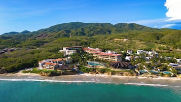 Un complejo turístico frente al mar con varias edificaciones, una playa y montañas al fondo.