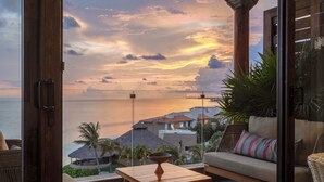 Un balcon avec vue sur l’océan au coucher du soleil, un canapé en osier, une table en bois et une porte vitrée.