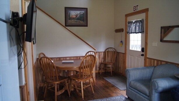 Uma sala de jantar com mesa e cadeiras de madeira, televisão fixada na parede e placa de saída acima de uma porta.