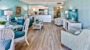 A living room with teal walls, a ceiling fan, a kitchen in the background, and a TV on a stand. - Emerald Isle 104 Beachfront|Updates Galore|Amazing SUNSET Views| 2 Bdrms (N. Redington Beach)