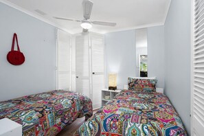 Une chambre avec deux lits, un ventilateur de plafond, un sac rouge accroché au mur et une fenêtre avec des stores.