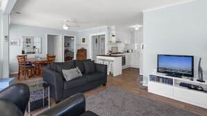 Ein modernes Wohnzimmer mit einer Couch, einem Essbereich mit Stühlen, einer Küche mit weißen Schränken und einem Flachbildfernseher auf einem Schrank.