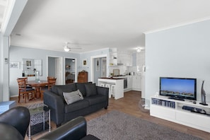 A modern living room with a sofa, a dining area with chairs, a kitchen with white cabinets, and a flat-screen TV on a stand.
