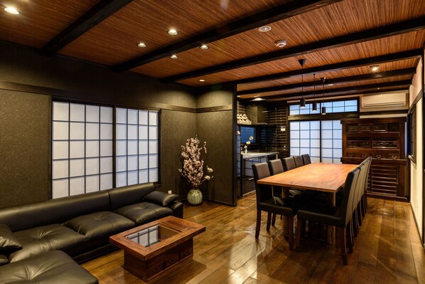 A modern interior with a wooden ceiling, a dining area with a wooden table and chairs, a black leather sofa, and a traditional Japanese-style window.