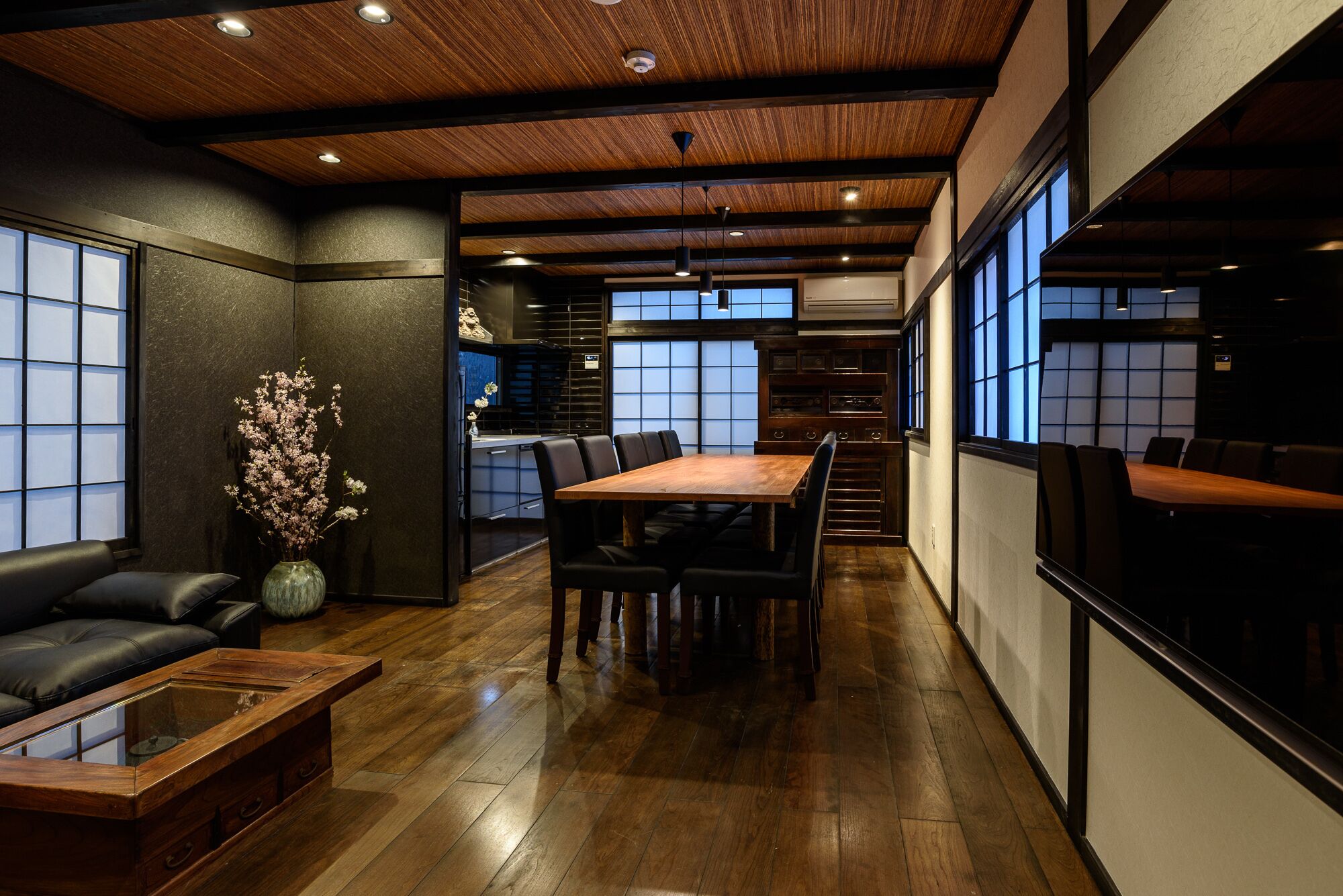 A modern interior with a wooden ceiling, a dining area with a wooden table and chairs, a sofa, and a vase of flowers.
