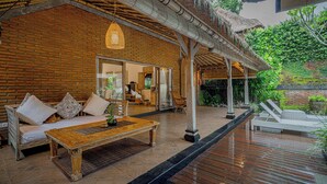 A spacious outdoor area with a wooden deck, a sofa with cushions, a hanging light fixture, and a thatched roof structure. - Charming Tropical Villa for Couple and Honeymooner (Gianyar)