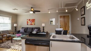 A compact kitchen with a dishwasher, a sink, and a ceiling fan. There is a dining area with a table and chairs, and a sofa in the background.