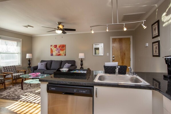 A compact kitchen with a dishwasher, a sink, and a ceiling fan. There is a dining area with a table and chairs, and a sofa in the background.