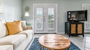 A living room with a beige sofa, a round wooden coffee table, a television on a black cabinet, and a door with white shutters. - Grand Caribbean East 111 * Fall Special Rates * Salt Water Vacations (Destin)