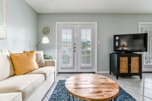 A living room with a beige sofa, a round wooden coffee table, a television on a black cabinet, and a door with white shutters.